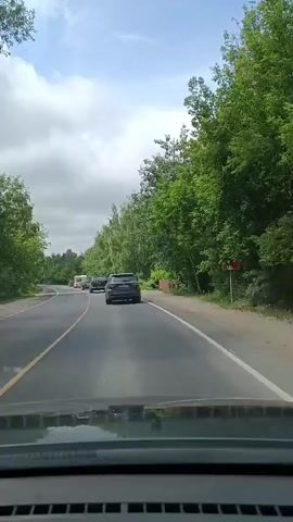 A rear view of a dark colored SUV driving on a paved road. The SUV has a license plate that reads "Y 754 OK 152". The road is flanked by trees and greenery on both sides. There are other vehicles ahead, including another SUV and a trailer. The sky is cloudy. The camera appears to be mounted inside another vehicle, looking out the windshield.