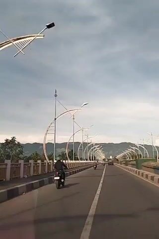 The video shows a view from a vehicle traveling on a highway overpass. The overpass is lined with decorative lampposts that have curved, arc-like structures. Several motorcycles are visible on the road, with riders in various clothing and helmets. The road has a white dividing line. In the background, there are mountains and a clear sky with some clouds. The scene appears to be during the daytime.