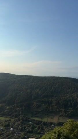 The video pans across a mountainous landscape bathed in sunlight. Initially, the view is of rolling, tree-covered hills under a clear blue sky with a few wisps of clouds. As the video progresses, the sun becomes more prominent, casting a warm glow and creating lens flares. The camera then focuses on a stone structure, possibly a bell tower or part of a building, with arched openings and decorative metalwork. The surrounding environment appears to be a rural area with vegetation and distant hills visible through the arches. The sun is visible in the sky throughout the latter half of the video.