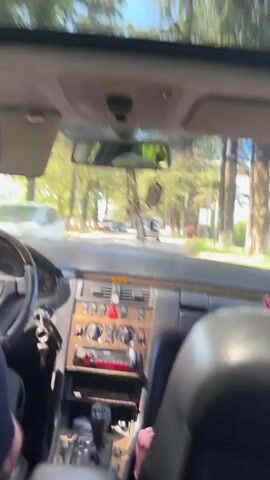 The video displays the interior view of a car, looking forward through the windshield, during a sunny daytime drive. The dashboard is dark with wood trim accents, featuring a multi-button center console, air vents, and a car radio with red backlighting. An automatic gear shifter with a wooden knob is visible, next to which a pack of Marlboro Red cigarettes is placed. Car keys are in the ignition.

The driver's left arm, wearing a dark short-sleeved shirt, is seen on the black steering wheel. The passenger's headrest is in the foreground on the right. A rearview mirror hangs several decorative charms, including a small framed icon. The word "AIRBAG" is visible on the passenger side of the dashboard.

Outside the car, a road lined with tall green trees stretches ahead. Other vehicles, including sedans and a white van, are visible in the distance. A circular road sign with a red border is partially seen on the right side of the road. The weather is bright and clear. Throughout the video, music with a male vocalist is playing in the background. The scene depicts a drive through Achadara, Abkhazia.