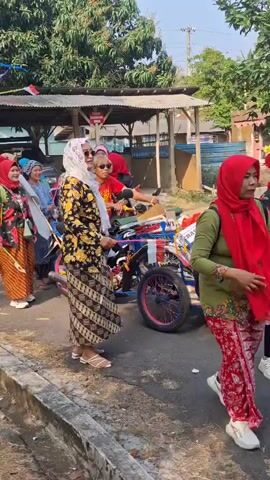 A group of people, mostly women wearing traditional Indonesian clothing and headscarves, are gathered outdoors. Some individuals are holding small Indonesian flags. A decorated motorcycle with a sidecar is present, adorned with colorful flags and streamers. The environment appears to be a street or pathway with buildings and trees visible in the background. The lighting suggests it is daytime. The people seem to be participating in a procession or event.