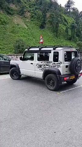 This is a short, horizontal video of a white four-door Suzuki Jimny parked on a paved road. The vehicle has black fender flares, black roof rails, and black wheels. There is a spare tire mounted on the rear door. The vehicle has a graphic decal on its side that says "JIMNY" and appears to be abstract and rocky in design. There are also some darker decals or graphics on the rear quarter panel. The vehicle is parked next to a hill with green trees and vegetation. There is a traffic sign visible on the hill behind the vehicle. The road surface is gray asphalt with some yellow markings. Another vehicle, partially visible, is parked to the left of the Jimny. The weather appears to be overcast.