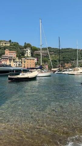 A drone shot pans across a harbor in a coastal town. Several boats, including a large yacht and a sailboat, are docked. The water is clear and shows the seabed. Buildings line the hillside behind the harbor, with lush green vegetation surrounding them. The sky is clear and blue. A person is briefly seen swimming in the water.