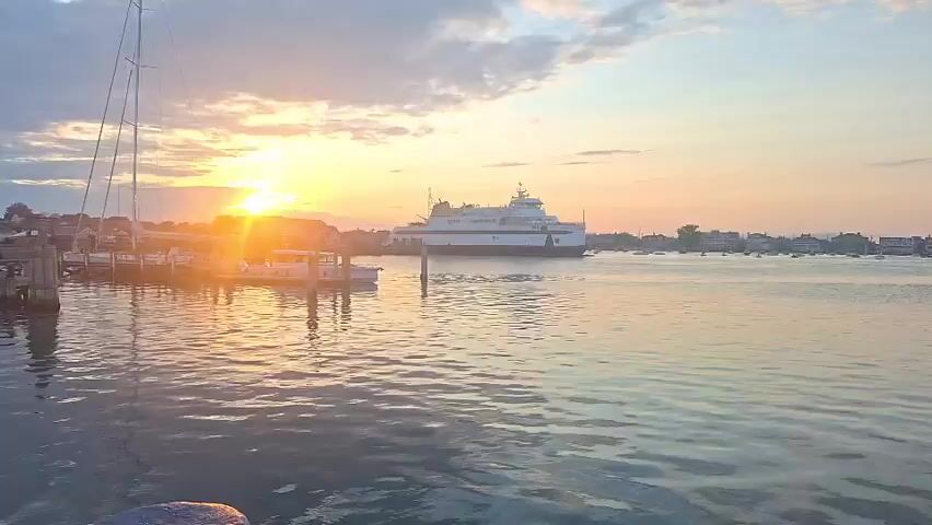 This video captures a serene harbor scene at sunrise. The sun, a bright orange orb, is low on the horizon, casting a warm glow across the water and illuminating the clouds. A large white ferry boat is docked in the distance, appearing as a prominent white structure against the colorful sky. To the left, a sailboat mast stands tall, its rigging silhouetted against the sunrise. Several smaller boats are moored in the foreground, gently bobbing on the water. The water itself reflects the sky's vibrant hues of orange, pink, and blue, creating a peaceful and picturesque atmosphere. Buildings line the distant shore, suggesting a coastal town or village. The overall impression is one of tranquility and the beauty of a new day beginning over the water.