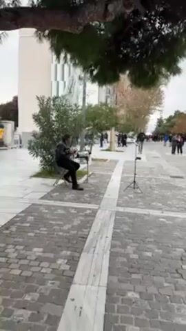 The video shows a man playing music on a paved outdoor walkway. The man sits on a folding chair, wearing a black jacket and dark pants. He is positioned to the left of the frame. He is playing an instrument, likely a keyboard of some kind, placed on a folding table in front of him. In front of the table, and towards the right of the frame, is a music stand on a tripod with what appears to be a donation box beneath it. The walkway is made of square paving stones with lighter-colored lines creating a grid pattern. In the background, there are blurred figures of people walking in the distance. There are buildings on either side of the walkway, including one with a large mural on its side. There are also trees around the walkway, some obscuring the view. The weather appears overcast. The video suggests a street performance or busking event.