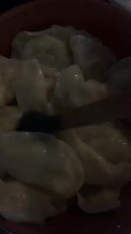 The video shows a close-up, overhead view of a bowl filled with pierogi. A dark utensil, possibly a fork or spoon, is seen moving amongst the pierogi. The pierogi are pale yellow and appear to be cooked, with visible crimped edges. The bowl itself is a reddish-brown color. The lighting is dim, making it difficult to discern finer details of the pierogi or the utensil.