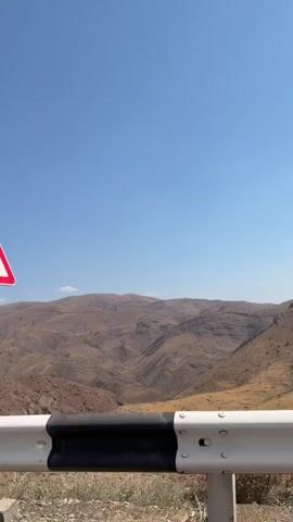 The video is a first-person perspective of a drive on a road. The road appears to be paved with asphalt and has white lane markings. In the foreground, there is a white metal guardrail with black and white reflective markings. Beyond the guardrail, the landscape is arid and mountainous, with dry vegetation and reddish-brown soil. The sky is clear blue with a few scattered white clouds. The video shows a curved road ahead with a road sign indicating a sharp turn to the right. The camera seems to be mounted on a moving vehicle, capturing the scenery as it passes. The audio in the video is the sound of the road noise from the vehicle.