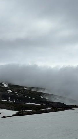 The video shows a snowy mountainous landscape on an overcast day. Thick clouds obscure the upper portions of the mountains and create a misty atmosphere. In the foreground, there is a snowy slope with a few tire tracks visible. Several people are scattered across the slope, some walking and others standing and taking pictures. The people are dressed in warm clothing, including jackets and hats, and some are holding walking sticks. The overall impression is of a cold, slightly eerie, but scenic environment.