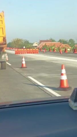 The video shows a view from inside a vehicle, looking out at a road under construction. Orange traffic cones and barriers are placed along the road, indicating a diversion or closure. A large yellow dump truck is visible on the left side of the frame, partially obscuring the view of the road ahead. The sky is clear and blue, and the sun appears to be shining. In the background, houses and trees can be seen, suggesting a rural or suburban setting. The overall impression is of a normal day with ongoing road work.