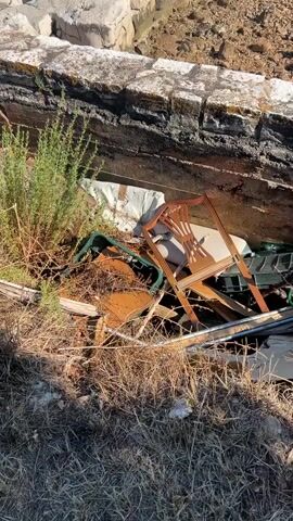 The video shows a pile of debris, including a wooden chair, green plastic crates, and various other discarded items. The debris is situated in a trench or eroded area next to a stone wall. Beyond the wall, there is a rocky shoreline with the sea visible. The ground is covered in dry grass and dirt. The overall impression is one of neglect and environmental damage. There are no people visible in the video, and no actions are being performed.