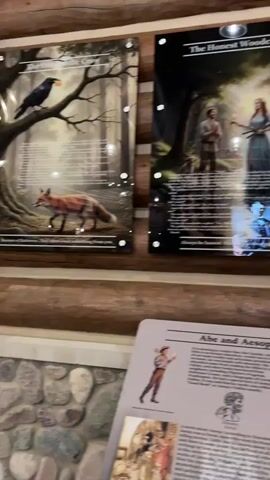 The video pans across a rustic log cabin interior, showcasing several framed exhibits mounted on the log walls. In the foreground, a sign titled "Abe and Aesop" is visible, featuring text and illustrations related to Abraham Lincoln and Aesop's Fables. To the left, an exhibit displays an illustration of a fox and text. To the right, two larger framed exhibits are mounted higher on the wall. The first depicts two figures in a forest setting with the title "The Honest Woodcutter," accompanied by extensive text. The second exhibit shows a scene with a stream and trees, also with accompanying text. The camera continues to pan, revealing large windows that look out onto an outdoor area with lighting and what appears to be outdoor seating. In the final shot, a book with the title "Outdoor Escapes" is resting on a surface in the foreground, with the log cabin walls and windows visible in the background. The lighting suggests it may be evening or a dimly lit interior.