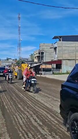 The video shows a street scene with construction activity. A large yellow steamroller is operating on a dirt road, compacting the surface. Several motorcycles are present, with people riding on them, navigating around the construction equipment. One motorcycle in the foreground has two passengers, a man driving and a woman and child behind him. Another motorcycle further back has a single rider. A black SUV is also visible, driving on the road. In the background, there are buildings and a tall communication tower. The sky is blue with some clouds. The road appears to be under construction or repair, as indicated by the presence of the steamroller and the loose dirt surface.