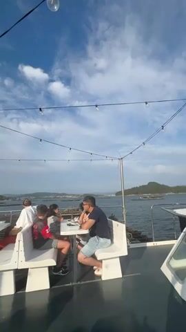 The video shows an outdoor seating area on what appears to be a boat or a pier, overlooking a body of water with a distant shoreline. Four individuals are seated at a white table. On the left, two people are sitting on a white bench. On the right, two people are seated at the table, one is a man wearing glasses and a dark blue t-shirt, and the other is a person wearing a red and black shirt. Several string lights are suspended overhead. The sky is blue with scattered clouds.