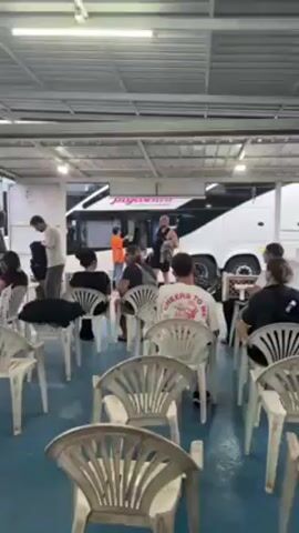 The video shows a gathering of people in what appears to be an outdoor waiting area. There are rows of white plastic chairs set up, some of which are occupied. Several white buses are parked nearby. People are either seated, standing, or moving around. Some individuals are looking at their phones. There are flags of various nations strung up in the background. The area is covered with a metal roof.
