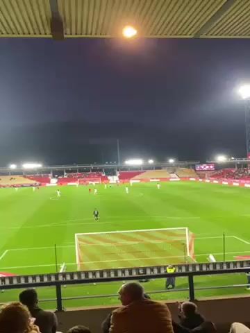 The video depicts a soccer game in progress from the perspective of a spectator seated in the stands. The stadium is brightly lit, with a green field and a mostly red seating area filled with spectators. The players, dressed in white and red uniforms, are actively engaged in the game. The camera captures the action from behind a goal net, with fans visible in the foreground, some sitting and others possibly standing.  Mountains can be seen in the background.  The video is shot at night.