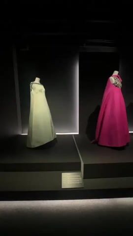The video pans across a dimly lit exhibition space, showcasing several mannequins dressed in formal gowns. The background is a dark, textured wall, with thin, vertical white light strips accentuating the display areas. In the foreground, on a raised black platform, a light green, floor-length gown with a cape is displayed on a mannequin. To its right, on a similar platform, a vibrant pink, floor-length gown with a detailed, embellished neckline and a cape is shown. The camera then moves to reveal a deep purple, floor-length gown with a halter neck, also on a mannequin. Further to the right, more mannequins are visible, one in a patterned, floor-length dress and another in a white, floor-length dress. The exhibition design features dark, stepped platforms with subtle LED lighting along the edges, creating a dramatic presentation for the garments. Informational plaques are placed on small stands in front of some of the displays. The overall ambiance is sophisticated and minimalist, focusing attention on the haute couture fashion.