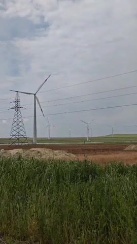 The video shows a landscape with multiple wind turbines and a power transmission tower. The turbines are white with red tips, standing tall in a field of dry, brown vegetation. Overhead power lines are visible, connecting the turbines to the electrical grid. In the foreground, there is a small mound of earth and patches of green grass. The sky is overcast with a mix of white and grey clouds. The camera is moving, likely from a passing vehicle.