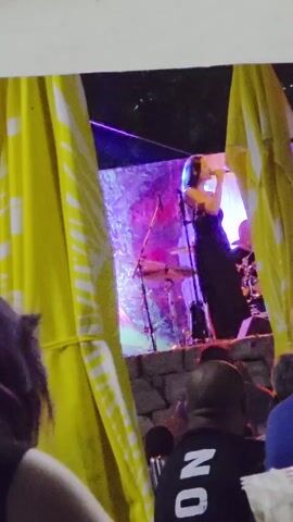 A woman sings into a microphone on a stage. She is wearing a dark dress with a strapless neckline. Behind her, a drum set is visible, as well as colorful stage lighting. Yellow curtains flank the stage. In the foreground, the heads and shoulders of audience members are visible. The scene appears to be an outdoor performance, possibly at night.