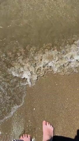 The video shows a top-down view of a person's bare feet standing on a sandy beach as a wave washes over them. The water is shallow and appears murky. The person is wearing a striped garment around their ankle. The wave recedes, leaving the sand wet. The video is 4 seconds long.