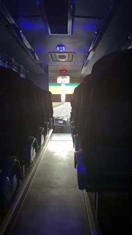 The media file shows the interior of a bus, looking forward towards the driver's cabin and the road ahead. Rows of dark seats line both sides of the aisle. Blue LED lights are visible along the ceiling of the bus, and a vent with a blue light is also present. A digital clock on the dashboard displays "20:26". Through the windshield, a paved road is visible, leading into a brightly lit area, possibly outdoors with natural light. A person is walking on the road in the distance. The scene appears to be set in the evening or night, given the interior lighting and the clock time. The context provided places the location in Шешенқара, Kazakhstan. No specific emotions are conveyed, and the overall impression is one of transit or waiting.