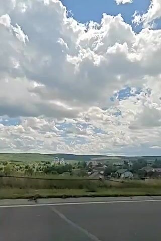 The video shows a view from a vehicle, likely a car, traveling along a road. The camera is pointed outwards, capturing a landscape on the right side. The landscape features rolling hills covered in green vegetation and a small village with houses. A bright blue sky with fluffy white and gray clouds dominates the upper portion of the frame. The road surface is visible in the foreground, appearing to be asphalt. The lighting suggests it is daytime, with the sun shining through breaks in the clouds. The movement of the vehicle is apparent from the slight blur and the way the landscape shifts in the frame. There are no people visible in the video.