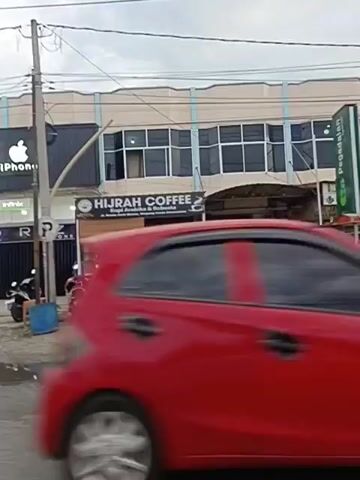 A street scene with several businesses, including a store labeled "iPhone" and "Raz Phone," and "Hijrah Coffee." Several motorbikes are parked or being ridden, and there are also a few cars. The sky is overcast. The video appears to be filmed from a moving vehicle.
