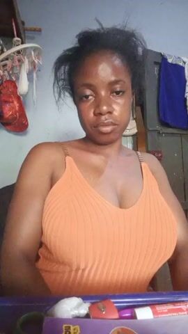 A woman with dark hair and skin is looking at the camera. She is wearing an orange tank top. She purses her lips. She is sitting at a desk with a purple box on it. There is a clothes hanger and a blue and white garment behind her.