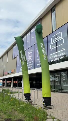 The video shows two inflatable tube men in front of a building. The tube men are green and yellow and are positioned side-by-side. They are blowing in the wind and appear to be advertising a store called "Tuinmeubel Shop". The building behind them has large windows with graphics of furniture, including a mattress and a crib. The text on the building reads "SHOPPING MALL". There are some metal railings and greenery in the foreground. The sky is cloudy.