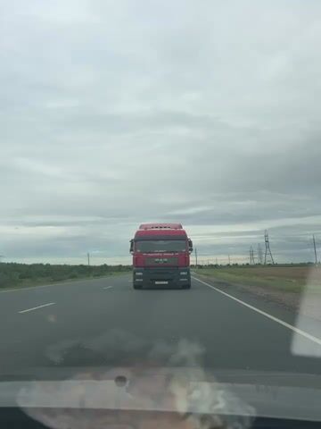 A red semi-truck is driving on a highway under a cloudy sky. The truck is in the center of the frame, approaching the camera. The highway has two lanes in each direction, and there is a grassy shoulder on either side. Power lines and fields are visible in the background. The video is shaky, suggesting it was recorded from a moving vehicle.