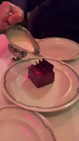 A person pours a white sauce from a silver gravy boat onto a chocolate dessert that is sitting on a white plate. The plate has a blue floral pattern around the edge. There are two more plates visible on the white tablecloth. The scene is lit with a pink light.