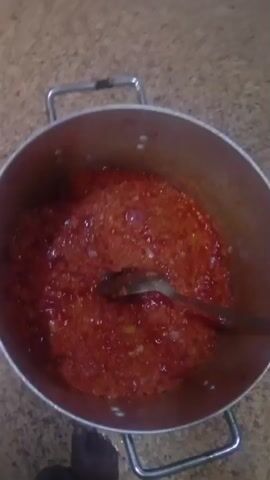 A close-up overhead shot shows a large metal pot filled with a red, chunky mixture. A metal spoon is stirring the contents of the pot. The pot has two silver handles. The background appears to be a countertop with a speckled texture. The lighting is dim, and the image quality is somewhat blurry.