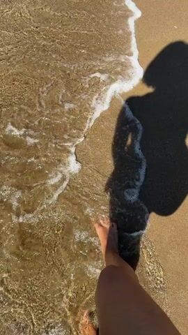 A first-person perspective video shows a person walking along a sandy beach where the water is shallow and clear. The shadow of the person is cast on the sand and in the water, showing a person with short hair carrying a bag over one shoulder. The waves gently lap at the person's feet. The sun is shining, creating a bright environment.