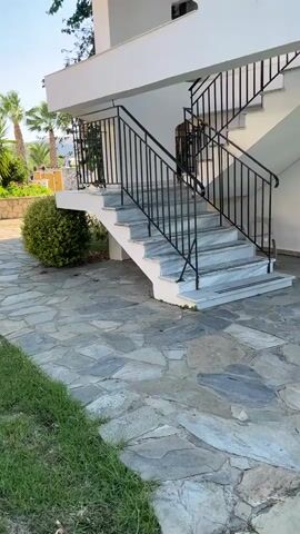 A first-person perspective video shows an outdoor setting with a paved walkway and green grass. To the right, a white building with a staircase leading up is visible, featuring black metal railings and marble-patterned steps. A small, rounded hedge is positioned next to the stairs. Further back, a landscaped area with palm trees and other greenery can be seen, alongside a low stone wall. To the left, a large tree with green leaves and a visible trunk stands in a grassy area, with part of a yellow building in the background. The sky is clear and blue, indicating daytime.