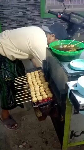 A person wearing a black baseball cap backwards, a light-colored long-sleeved shirt, and a patterned sarong is standing next to a food cart. They are bent over, tending to a grill with numerous skewers of round, golden-brown meatballs cooking over red coals. To their right, on top of the cart, is a large green bowl filled with broth and more meatballs on skewers. Behind the person and the cart is a motorcycle. The environment appears to be outdoors at night, with a patterned tile wall in the background. The person is actively engaged in cooking, possibly fanning the coals or adjusting the meatballs.
