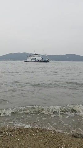 A white ferry boat is in the middle of a body of water. The water is gray and choppy with small waves crashing on a rocky shore in the foreground. The sky is overcast and gray. Hills are visible in the background.
