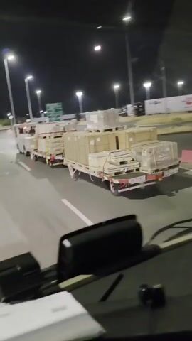 A camera mounted inside a vehicle captures a nighttime view through the windshield and side mirror. The vehicle appears to be a truck, as indicated by the dashboard and the driver's side mirror. The truck is moving on a paved road, possibly an airport tarmac or a logistics yard, as suggested by the presence of other vehicles and infrastructure like lighting poles and signs. In the foreground, the truck's dashboard is visible. In the middle ground, several flatbed trailers loaded with boxes are being towed by small utility vehicles. The boxes are stacked and secured with straps. The trailers and utility vehicles are moving in the same direction as the camera vehicle. In the background, large semi-trailer trucks and buildings are visible. The lighting conditions are dim, with artificial lights illuminating the scene. The moon is also visible in the dark sky. The overall scene depicts a logistics operation, likely at an airport or a large distribution center, during nighttime hours.