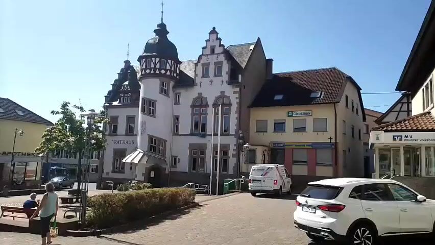 The video opens on a bright, sunny day in what appears to be a European town square. In the foreground, a white SUV is parked facing the right side of the frame. To the left, a cobblestone pathway leads towards a multi-story building with a distinctive turret and ornate architecture, labeled "RATHAUS," which translates to "Town Hall." Adjacent to the Town Hall is a row of businesses with storefronts. One of these businesses has a sign that says "Sparkasse." Further to the right, a modern building with a glass entrance and banking signage ("Volksbank") is visible. The camera pans slightly to the right, showing more of the banking establishment and the street. A white van with "KLEMPNER" written on it is parked in front of the Town Hall. In the lower left corner, a person is sitting on a bench, and a small tree is present. The overall impression is a quiet, well-maintained town center under a clear blue sky.