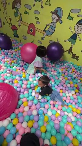Two young children are playing in a ball pit. The pit is filled with pastel-colored plastic balls and a few larger exercise balls. The wall behind the pit has a yellow background with cartoon drawings of children playing on seesaws and other playground equipment. The children are both seated in the ball pit, and the boy is initially prone before sitting up. The girl is interacting with a large, purple exercise ball.