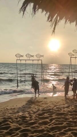 The scene is a beach at sunset. The sun is bright and low, creating a glare on the water. Several people are present on the beach. One man is swinging on a swing set that appears to be partially submerged in the water. Another person is playing in the water nearby. Additional people stand on the beach. Decorative fish silhouettes adorn the top of the swing set. The beach sand is textured with footprints and patterns. There is a structure with a thatched roof visible in the foreground.