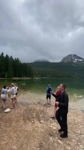 The video captures a group of people gathered at the edge of a tranquil mountain lake under an overcast sky. In the initial frame, a man dressed in a black suit and black shoes walks towards the water, holding a mobile phone in his right hand. To his left, two women in light-colored tops and shorts stand near the water. As the camera subtly pans right, more individuals come into view. A child in a blue top and shorts stands by the water's edge, while another child in a bright green top holds a stick, looking out at the lake. A woman in a red jacket and patterned shorts, and another woman in jeans and a maroon top, are also part of the group, observing the scenic view.

The lake itself features remarkably clear, green-tinged water, allowing the sandy bottom to be visible in the shallows. The background is dominated by lush green pine forests that extend up the slopes of large mountains, whose peaks are partially covered with patches of snow. The sky is filled with heavy, grey clouds, suggesting an overcast day. A small sign, whose text is illegible, is partially submerged in the shallow water close to the shore. The overall impression is one of people enjoying the serene and natural beauty of a high-altitude lake environment.
