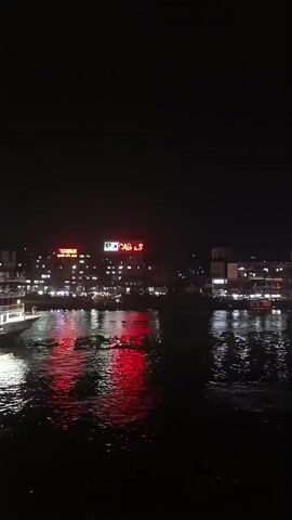 The video shows a night view of a river with a large, lighted boat passing by. The boat appears to be a multi-decked vessel with lights illuminating its structure. On the opposite bank of the river, there are buildings with neon signs, one of which clearly reads "CABLES". The lights from the buildings and the boat are reflected on the dark water, creating shimmering red and white patterns. The scene is generally dark with the lights serving as the primary source of illumination. The camera appears to be positioned on another vessel or structure, as part of a railing is visible in the foreground.