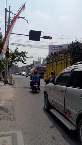 A view from a sidewalk looking at a street with moderate traffic. A traffic light is red. A silver car is parked on the right side of the street. A yellow truck loaded with sugar cane is in the intersection. Several motorcycles are in the street. A person on a motorcycle is wearing a backpack. A flag with red and white stripes is displayed on a pole on the left. The sky is clear and bright.