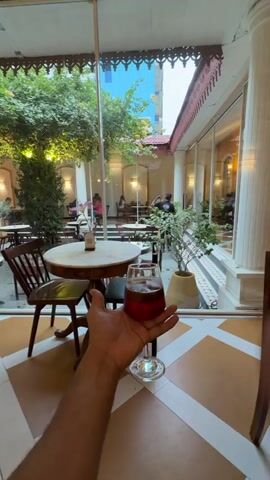 The video shows a first-person perspective of someone holding a glass of red liquid. In the foreground, there is a wooden table and chair. The background is a courtyard with more tables and chairs, greenery, and people sitting at the tables. The courtyard is surrounded by a building with large windows and decorative architectural elements. The overall impression is a cafe or restaurant with an outdoor seating area.