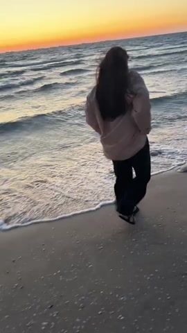 A young woman with long, dark hair is filmed from behind, standing on a sandy beach at sunset. She is wearing a light-colored top and dark pants. The ocean stretches out before her, with gentle waves lapping at the shore. She raises her arms and appears to be stretching or adjusting her hair, her head slightly tilted. The sky is a warm orange and yellow, indicating either sunrise or sunset. The overall atmosphere is peaceful and serene.