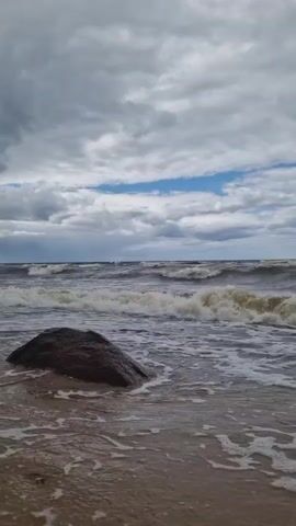 A video shows a turbulent ocean under a cloudy sky. Waves crash onto a sandy shore, with a large rock visible in the foreground. The water is dark and choppy, with white foam churning. The sky is filled with gray clouds, but patches of blue sky are visible. The overall mood is one of wild, untamed nature.