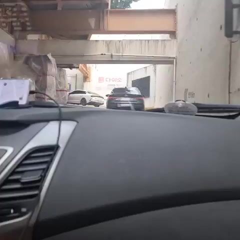 The video shows the interior of a car, looking out through the windshield at traffic. Two cars are visible ahead, one black and one white. The black car is closer and in the center of the frame, while the white car is further back and to the left. There is a building with a sign that says "다이소" (Daiso) in the background. The road appears to be under an overpass or bridge, with exposed beams and concrete visible above. The car's dashboard is in the foreground, with a phone mounted on the left side. The scene appears to be a street in an urban area, likely in South Korea due to the Korean signage.