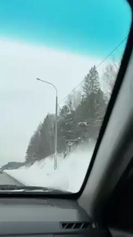 The video shows a car driving on a snow-covered road. On the right-hand side of the road, there is a small hill covered in snow and trees. The sky is overcast. The car is moving at a steady speed. The camera is located inside the car, capturing the view through the windshield. There is a road sign visible in the distance. Music is playing.