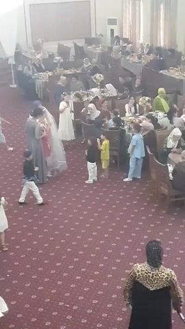The video shows a formal indoor gathering, possibly a wedding reception, with guests seated at tables in the background. In the foreground, a bride, wearing a veil and a white dress, is surrounded by several people. A woman in a grey dress stands to the bride's left, and another woman in a white garment stands to the bride's right, holding what appears to be a bouquet. A group of children are present, some observing the bride and her companions. One child, dressed in white, stands to the left of the bride. Another child, in a blue shirt, is to the right, near the woman in white. A child in a black shirt and white pants is positioned in front of the bride, facing away from the camera. The floor is covered with a red carpet featuring a repeating pattern. The lighting appears to be artificial, casting a warm glow over the scene.