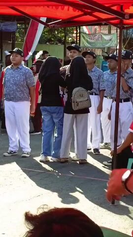The video shows a group of young people standing under a red canopy. Several individuals are wearing matching uniforms that include white pants, patterned shirts, and dark caps. Some are holding what appear to be flags or banners. In the foreground, a person's hand and arm are visible, with a red watch on their wrist, and they are pointing towards the group. Two individuals in the center of the frame are interacting, one wearing a black hijab and the other a dark top and a checkered backpack. Another person in the background is wearing a red and gold uniform. The environment appears to be outdoors, with sunlight casting shadows on the ground. There are also other people and structures visible in the background.
