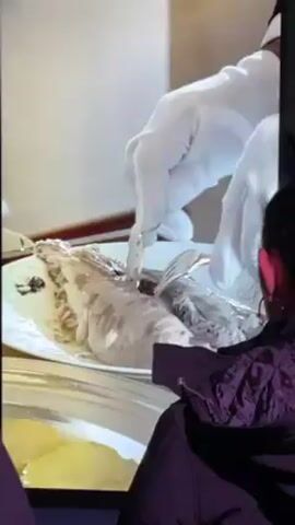A woman in a purple jacket is watching a video of gloved hands meticulously filleting a fish. The fish sits on a white plate. The gloved hands use a knife and fork to remove the bones. The woman is an observer of the video and does not interact with it.